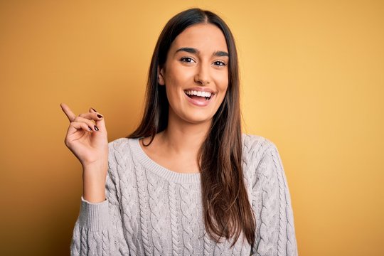 Young beautiful brunette woman wearing white casual sweater over yellow background with a big smile on face, pointing with hand and finger to the side looking at the camera.