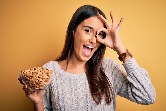 Young beautiful brunette woman holding bowl with peanuts over isolated yellow background with happy face smiling doing ok sign with hand on eye looking through fingers
