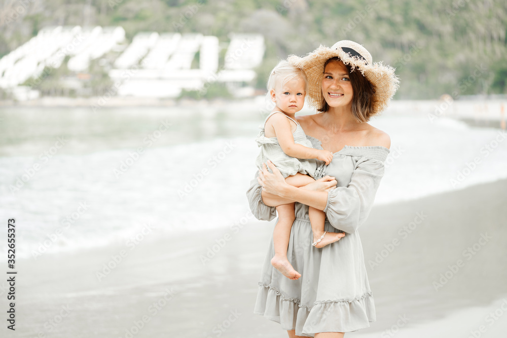 Wall mural young happy mother in a straw hat holds a blonde girl in the arms of her child and looks at the came