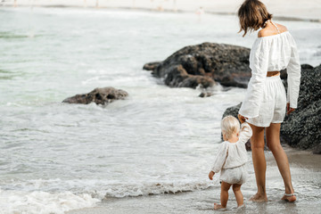 Young slender mom and her child on the beach near the water in white stylish clothes. Childhood and...