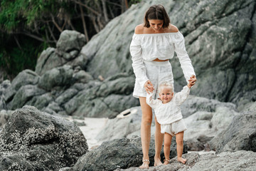 Portrait of a young mom and her child walking in white clothes barefoot on the beach stones....