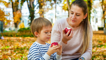 Portrait of little todler boy with mother eating red apples in autumn park
