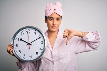 Young beautiful brunette woman wearing pajama and sleep mask holding big clock with angry face,...
