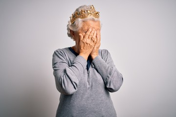 Senior beautiful grey-haired woman wearing golden queen crown over white background with sad expression covering face with hands while crying. Depression concept.