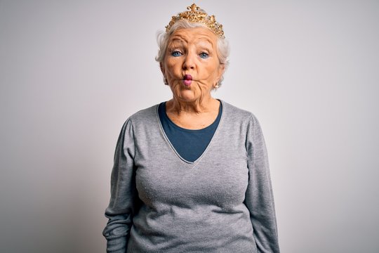 Senior beautiful grey-haired woman wearing golden queen crown over white background making fish face with lips, crazy and comical gesture. Funny expression.