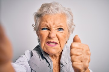 Senior beautiful grey-haired woman making selfie by camera over isolated white background annoyed and frustrated shouting with anger, crazy and yelling with raised hand, anger concept
