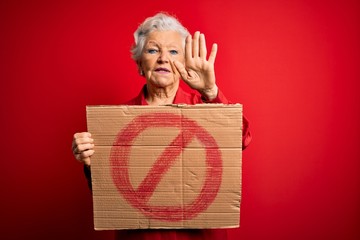 Senior beautiful grey-haired woman holding banner with prohibited signal over red background with open hand doing stop sign with serious and confident expression, defense gesture
