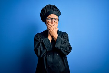 Senior beautiful grey-haired chef woman wearing cooker uniform and hat over blue background shocked covering mouth with hands for mistake. Secret concept.