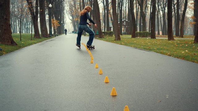 Shooting from below of experienced bearded man with long hair roller skating, skillfully skating and doing various complex tricks and turns between training cones in city park in damp spring weather.