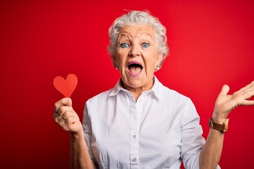 Senior beautiful woman holding paper heart standing over isolated red background very happy and...