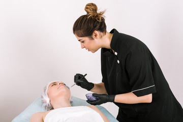 Obraz na płótnie Canvas Girl cosmetologist isolated from white background in medical clothes and protective disposable hygienic gloves with a brush puts treatment substance on face of female patient lying on table.