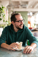 Vertical frame portrait of handsome real guy with eyeglasses beard and casual outfit sitting at bar counter with cup of coffee in his hand looks away. Developmental brain activity concept