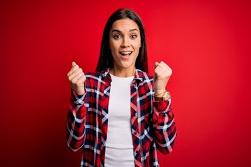 Young beautiful brunette woman wearing casual shirt standing over isolated red background celebrating surprised and amazed for success with arms raised and open eyes. Winner concept.