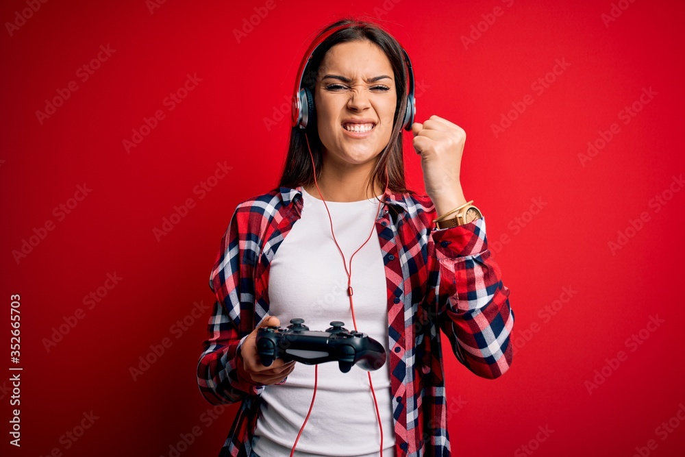 Wall mural young beautiful brunette gamer woman playing video game using joystick and headphones annoyed and fr