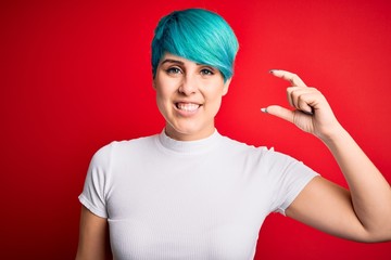 Young beautiful woman with blue fashion hair wearing casual t-shirt over red background smiling and confident gesturing with hand doing small size sign with fingers looking and the camera. Measure