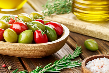 Green and red big olives in a wooden plate. Olive oil, rosemary, thyme, salt, pepper