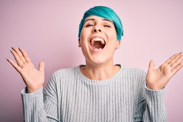 Young beautiful woman with blue fashion hair wearing casual sweater over pink background celebrating mad and crazy for success with arms raised and closed eyes screaming excited. Winner concept