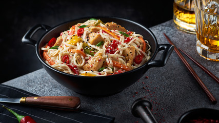 Wok with noodles, chicken and vegetables in a black asian bowl. Whisky, stylish chopsticks, chili pepper. Grey surface, black background. Asian style, chinese cuisine