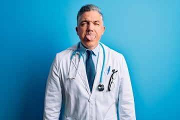 Middle age handsome grey-haired doctor man wearing coat and blue stethoscope sticking tongue out happy with funny expression. Emotion concept.