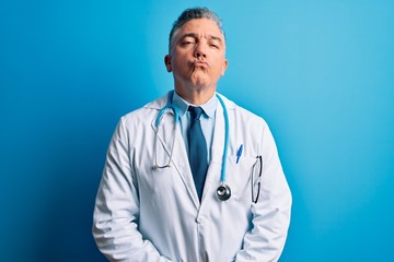 Middle age handsome grey-haired doctor man wearing coat and blue stethoscope looking at the camera blowing a kiss on air being lovely and sexy. Love expression.