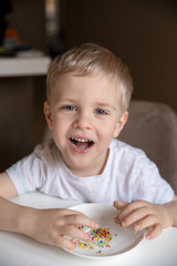 Smiling little boy with blond hair in a white T-shirt eats sweets. The boy is having fun, he eats sweets with his hands.