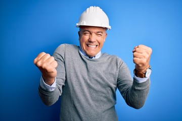 Middle age handsome grey-haired engineer man wearing safety helmet over blue background very happy and excited doing winner gesture with arms raised, smiling and screaming for success. Celebration