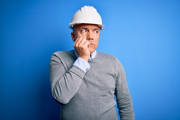Middle age handsome grey-haired engineer man wearing safety helmet over blue background looking stressed and nervous with hands on mouth biting nails. Anxiety problem.