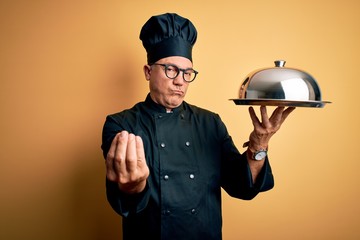 Middle age handsome grey-haired waiter man wearing cooker uniform and hat holding tray doing money gesture with hands, asking for salary payment, millionaire business - obrazy, fototapety, plakaty