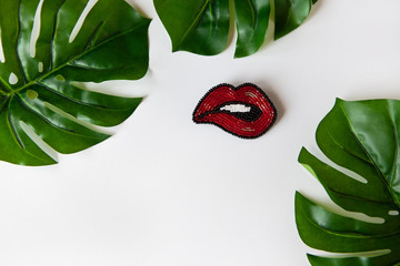 Fashionable brooch in shape of lips from Japanese beads, green leaves of Monstera on white...