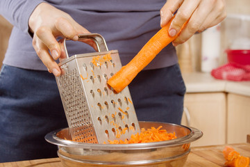 Dłonie kobiety trzymają marchewkę i ścierają ją na tarce kuchennej.
