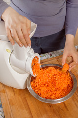 Maszynka elektryczna z nakładką do szatkowania warzyw. Kobieta w kuchni szatkuje marchew na drobne kawałki.