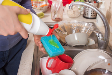 Kobieca dłoń trzyma gąbkę do mycia naczyń na którą nalewa z białej butelki płyn do mycia naczyń. Na drugim planie zlew zapełniony brudnymi naczyniami.