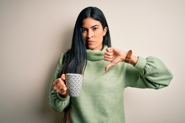 Young beautiful hispanic woman drinking a cup of hot coffee over isolated background with angry face, negative sign showing dislike with thumbs down, rejection concept