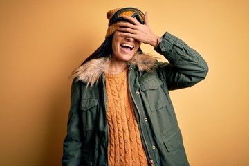 Young beautiful hispanic woman wearing ski glasses and coat for winter weather smiling and laughing with hand on face covering eyes for surprise. Blind concept.