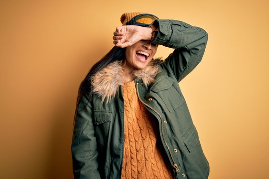 Young beautiful hispanic woman wearing ski glasses and coat for winter weather covering eyes with arm smiling cheerful and funny. Blind concept.