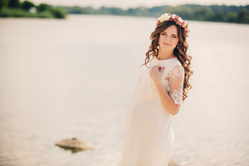 Fototapeta na wymiar beautiful young pregnant woman is wearing white dress and floral wreath near the lake, summer time, pregnant girl. concept of motherhood