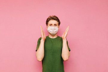 Portrait of a shocked young man in a white gauze mask standing on a pink background and with raised hands emotionally looking into the camera. Coronavirus pandemic. Quarantine. covid-19.