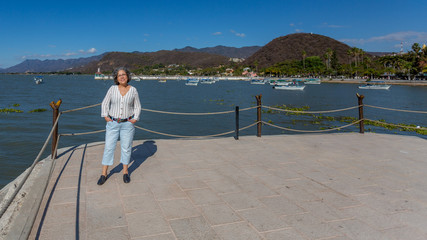 Fototapeta na wymiar Smiling tourist with casual clothes posing on the boardwalk with the lake with motorboats, the pier and the mountains in the background, sunny day with a clear blue sky in Chapala, Jalisco, Mexico