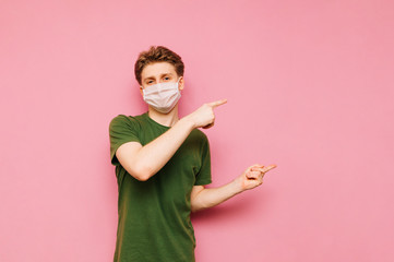 Positive young man in a green t-shirt on a white protective mask looks at the camera and points his fingers to the side on the copy space. Coronavirus pandemic. Quarantine. covid-19.pink background