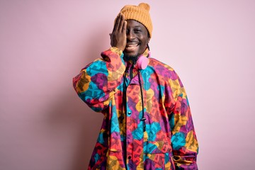 Young handsome african american man wearing colorful coat and cap over pink background covering one eye with hand, confident smile on face and surprise emotion.