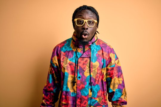 Handsome african american man wearing colorful coat and glasses over yellow background afraid and shocked with surprise expression, fear and excited face.