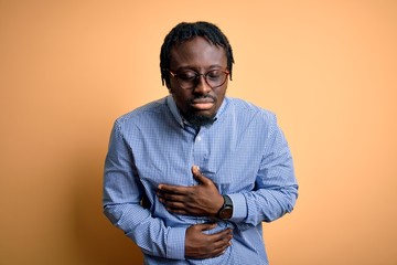 Young handsome african american man wearing shirt and glasses over yellow background with hand on stomach because indigestion, painful illness feeling unwell. Ache concept.