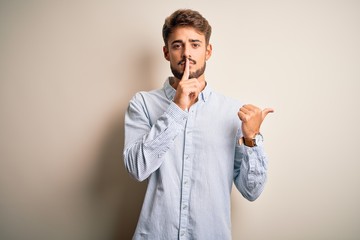 Young handsome man with beard wearing striped shirt standing over white background asking to be quiet with finger on lips pointing with hand to the side. Silence and secret concept.