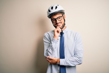 Young businessman wearing glasses and bike helmet standing over isolated white bakground looking confident at the camera with smile with crossed arms and hand raised on chin. Thinking positive.
