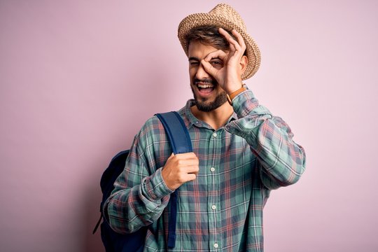 Young tourist man on vacation wearing backpack and summer hat over pink background with happy face smiling doing ok sign with hand on eye looking through fingers