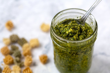 Jar of vegan basil pesto with a spoon and dried pasta scattered around the jar on a white and gray marble countertop