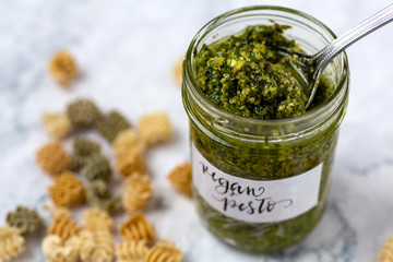 Jar of vegan basil pesto with a spoon and dried pasta scattered around the jar on a white and gray marble countertop