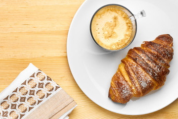 Top view on a breakfast consisting of a cup of coffee, a croissant on a wooden table with copy space. Serviette near the white plate with snack. 