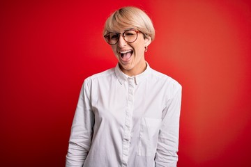 Young blonde business woman with short hair wearing glasses over red background winking looking at...