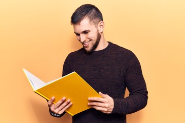 Young handsome man reading book looking positive and happy standing and smiling with a confident smile showing teeth